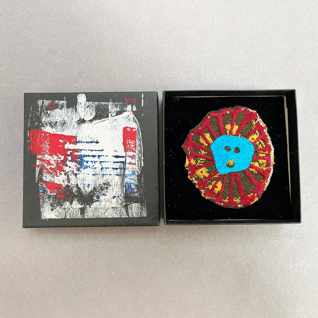 A machine embroidered brooch depicting a blue and burgundy skull by Michael Brennand -Wood next to it's own unique hand-painted presentation box.