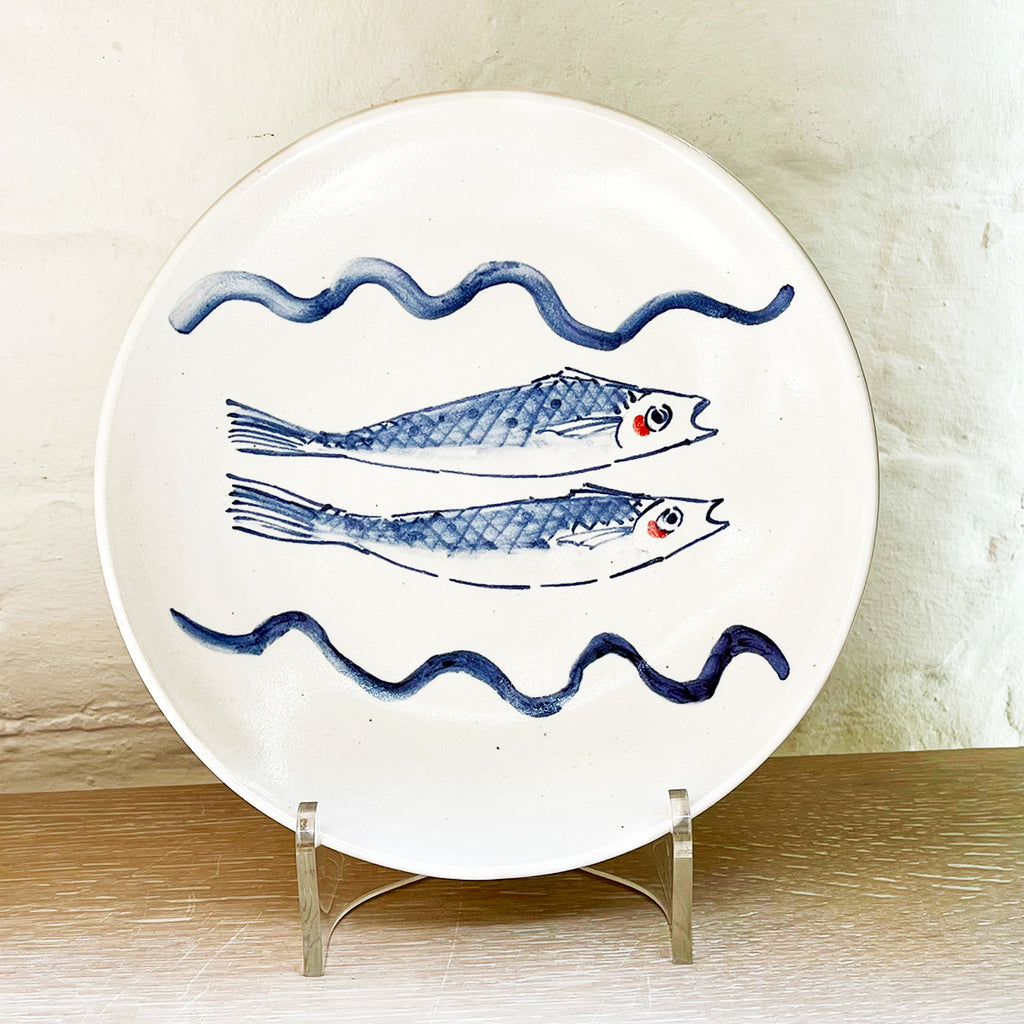 A beautiful round serving dish made from white-glazed stoneware and hand decorated with two cobalt blue fish. It is hown on a stand (not included) at the Bluecoat Display Centre, home of contemporary ceramics and gifts