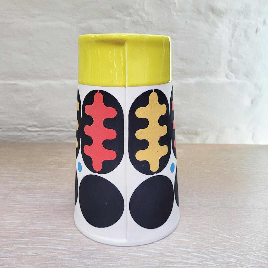 rear view of the Large Flat Top Cone Vase decorated using stencils and slip colours. With a distinctively mid-centurary modern feel about it - this piece is predominently white background and navy with red, blue and peach accents and a yellow top section. This is by Maree and Paul Allitt and is at The Bluecoat Display Centre Liverpool