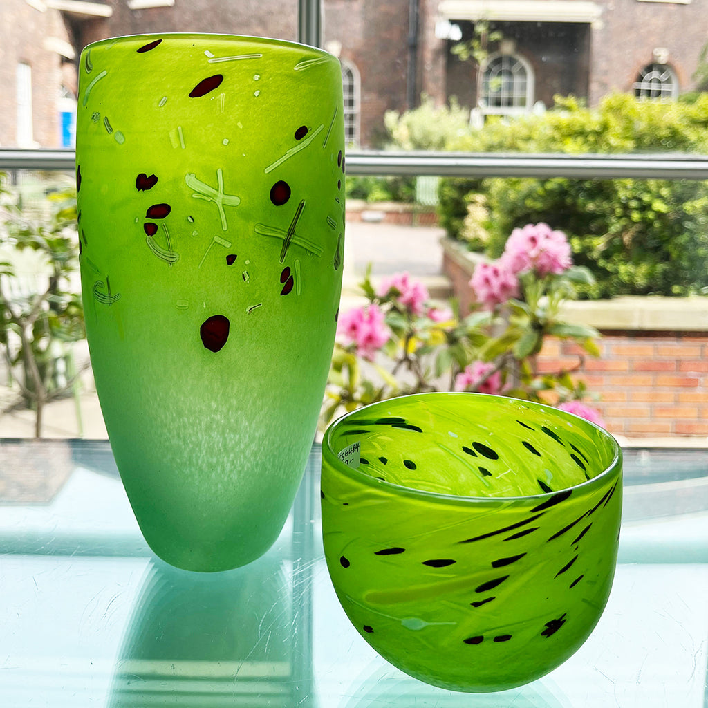 A medium cone vase next to a small bowl by contemporary glass artist Will Shakspeare. Pieces inspired by the landscape of the Somerset Levels and  predominantly green in colour with accents in red, black and white. Shown at the Bluecoat Display Centre window