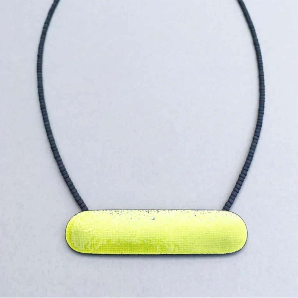 Close up photo of bright acid yellow necklace with a long horizontal textured shape partly enamelled strung on cable with tiny gunmetal colour glass seed beads by Annabet Wyndham