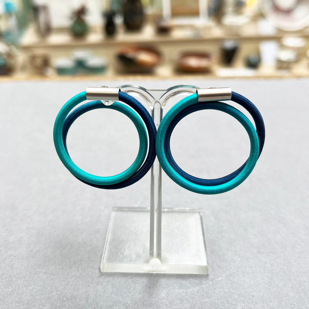 contemporary blue green double loop earstuds are the perfect way to add a pop of colour to any outfit. In silver and hand dyed elastic by Gilly Langton. Photographed on a stand in the Bluecoat Display Centre