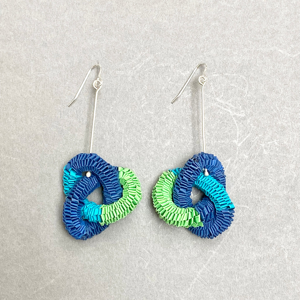 knotted paper earrings in tones of light blue, dark blue and green twisted throughout each other on grey background by Rike Lenzing