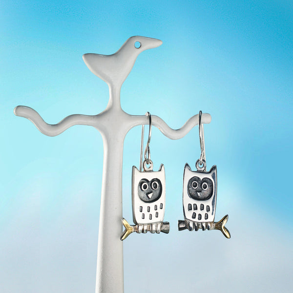 Silver owl earrings by Contemporary Irish jeweller Alan Ardiff, hanging from a branch against a blue gradient background